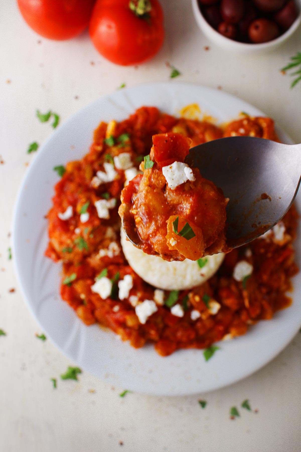 A spoon held in mid-air with a juicy shrimp on it, hovering above a plate of traditional shrimp saganaki.
