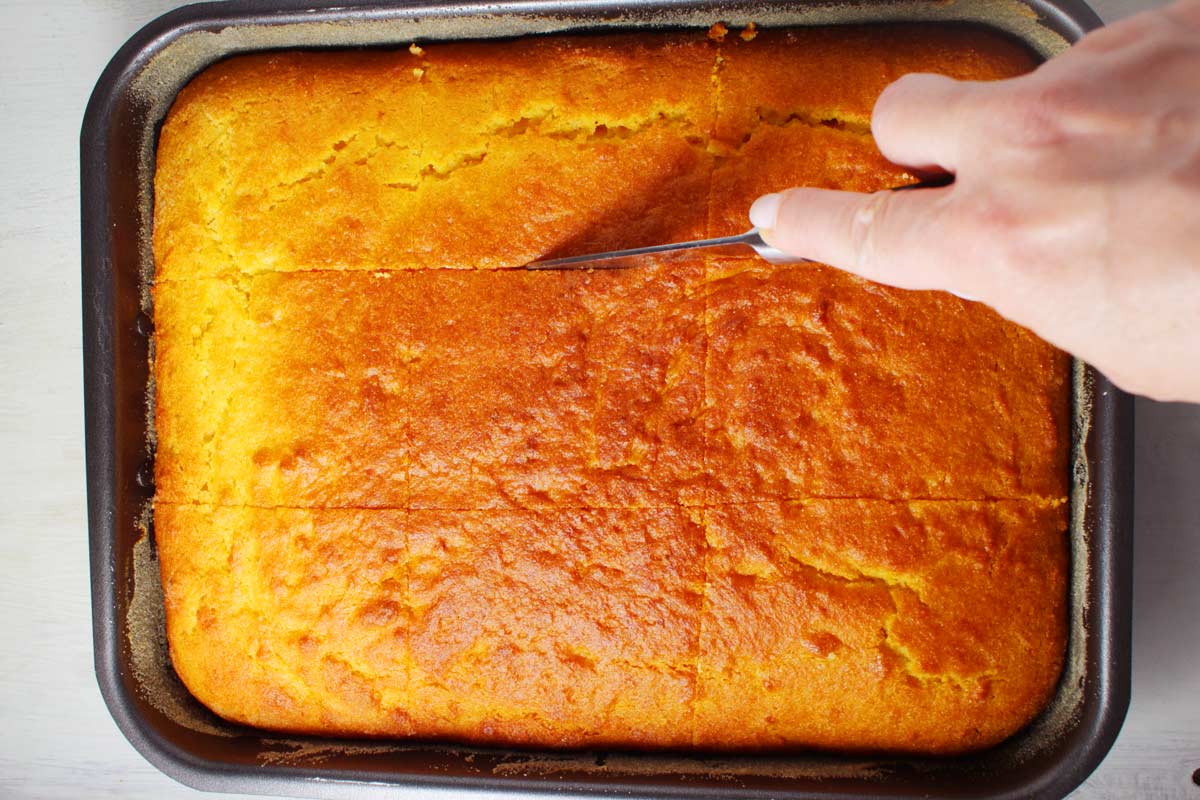 A hand tht is holding a knife and cutting pieces over a baking pan with revani.