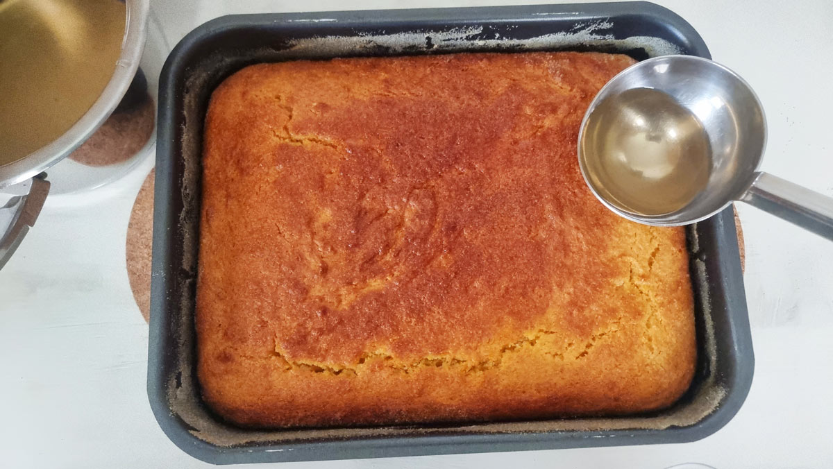 A baking pan with revani and a hand that is laddling syrup over it.