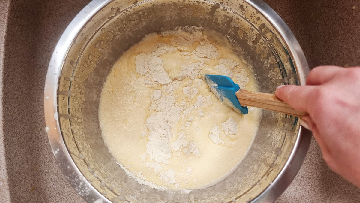 A mixing bowl with a combination of dry and wet ingredients and a hand that is holding a spatula.