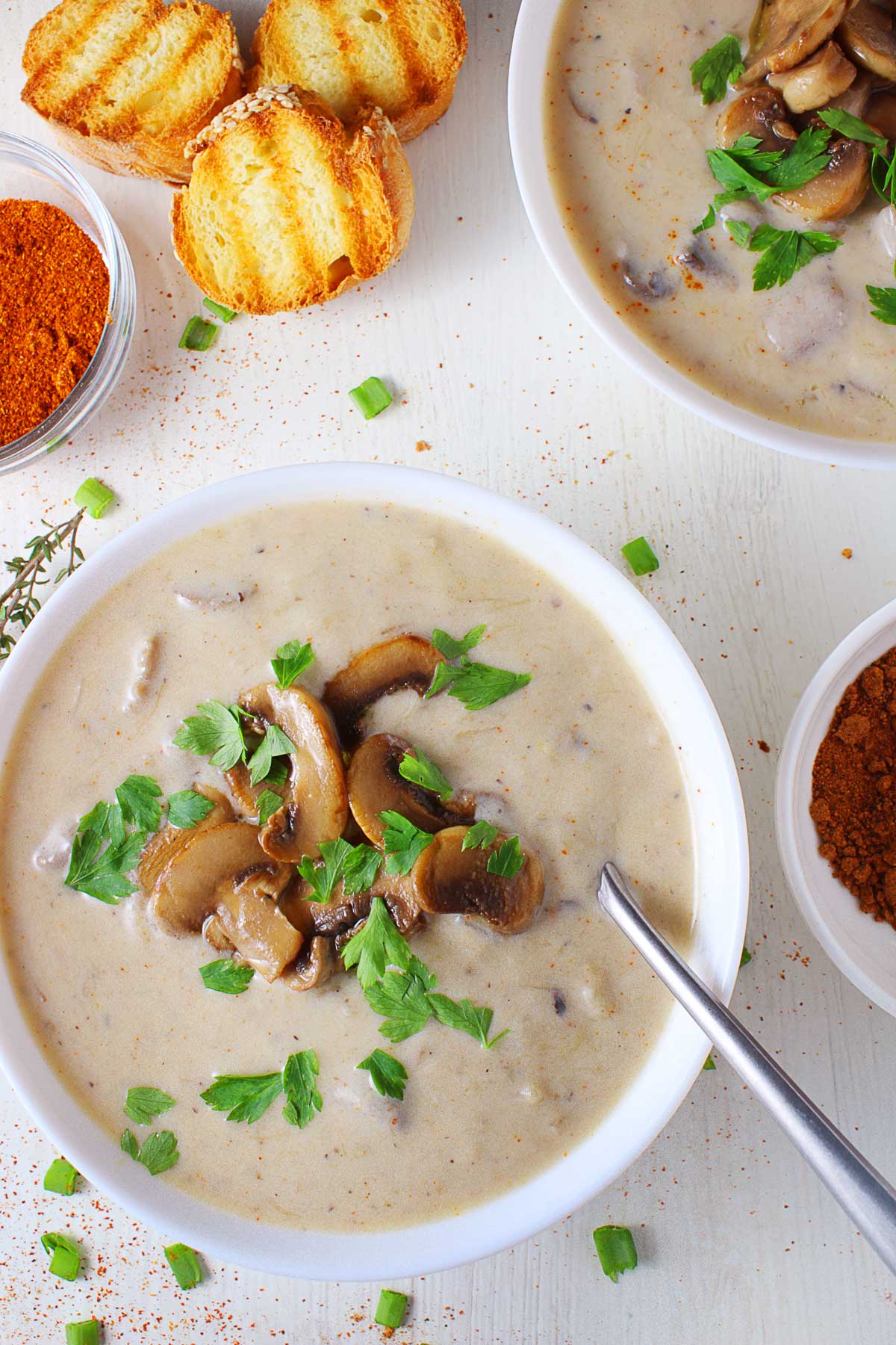 Two white bows with Mediterranean mushroom and leek soup surrounded by bread and small bowls with spices.