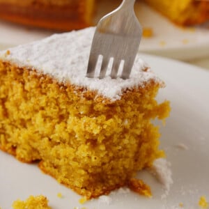 The featured image of the post "Greek vegan cake - Fanouropita. A piece of cake on a white plate with a fork ready to cut through another portion.