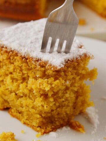 The featured image of the post "Greek vegan cake - Fanouropita. A piece of cake on a white plate with a fork ready to cut through another portion.