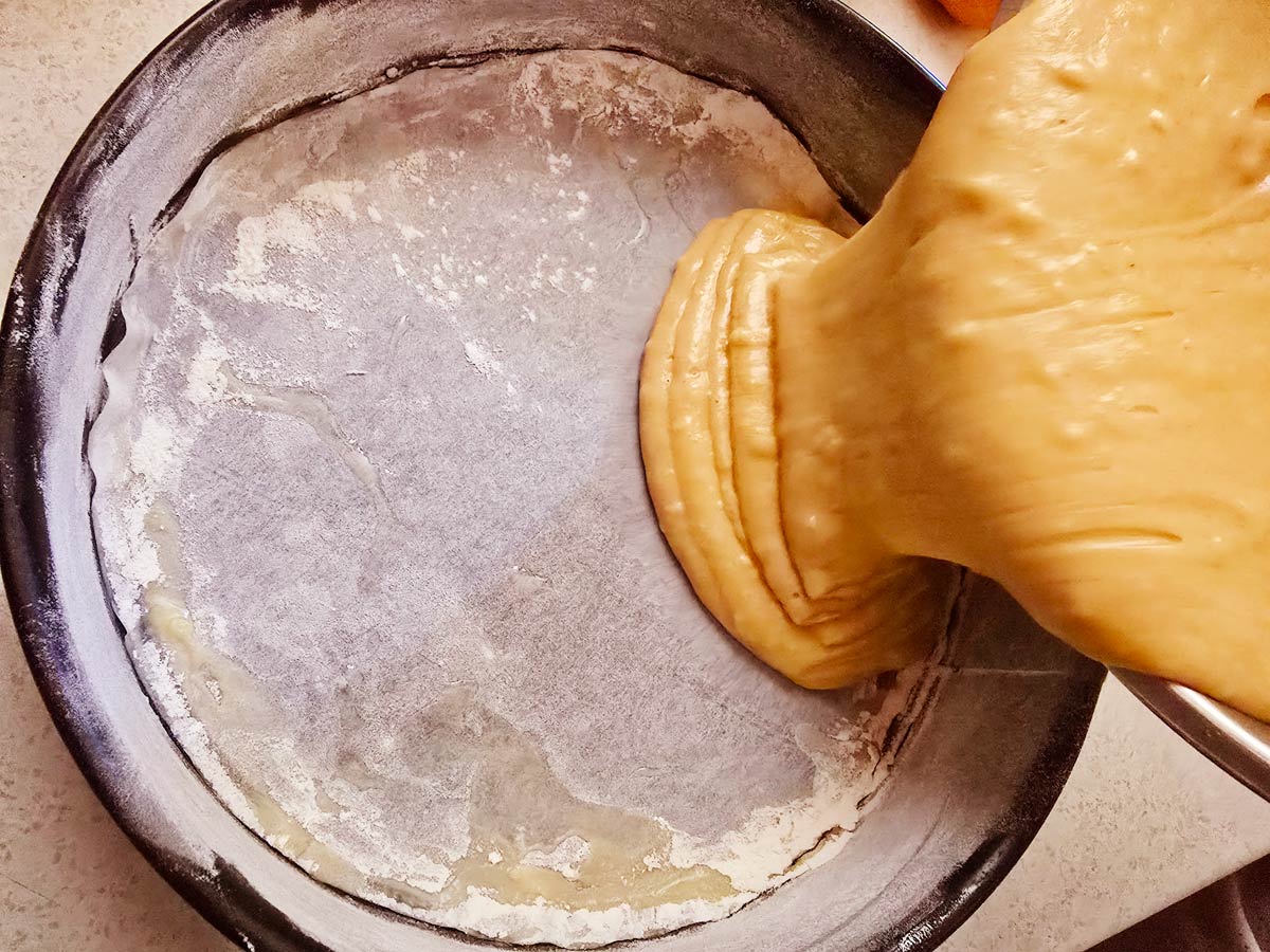 A batter for making Greek vegan cake is pouring into a black round baking pan.
